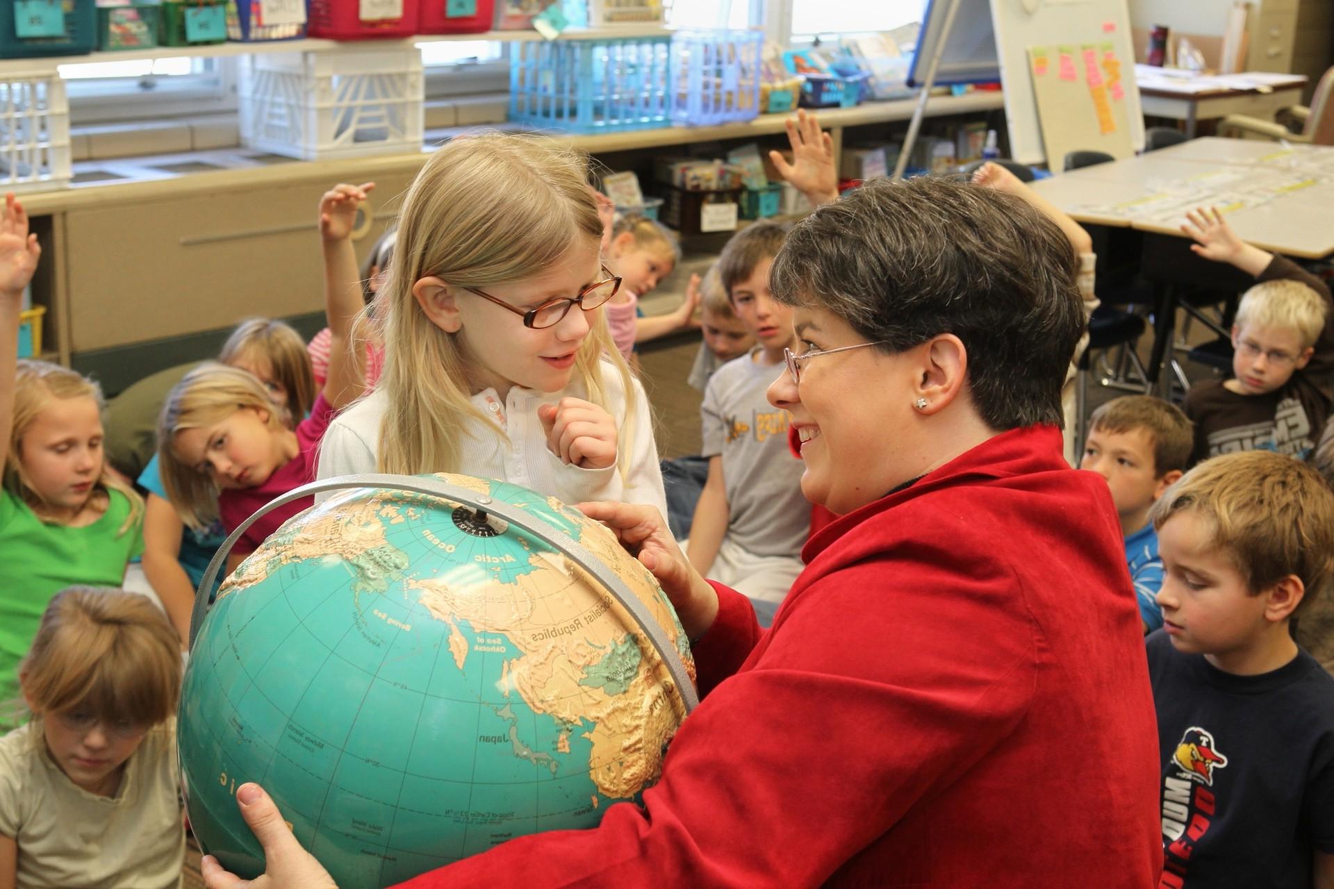 A female 世界语言教师 points out a country on a globe to a little girl as the rest of the class sits on the floor around them.